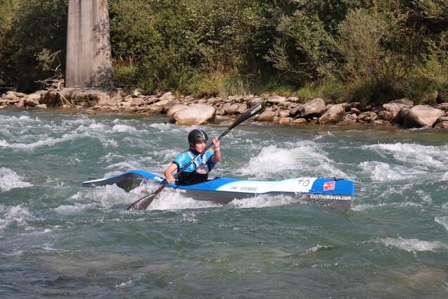 Paddler in einem blauen Kajak manövriert durch rauschendes Wasser, umgeben von Felsen und grüner Vegetation.
