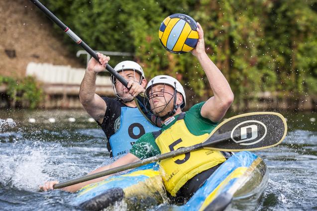 Zwei Kanuten in einer aufregenden Spielsituation, einer hält einen Wasserball, während der andere mit dem Paddel agiert.