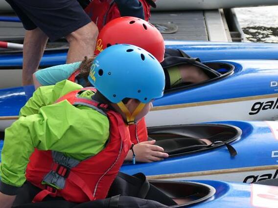 Kinder in Kayaks mit Schwimmwesten und Helmen, bereit zum Paddeln an einem Steg am Wasser.