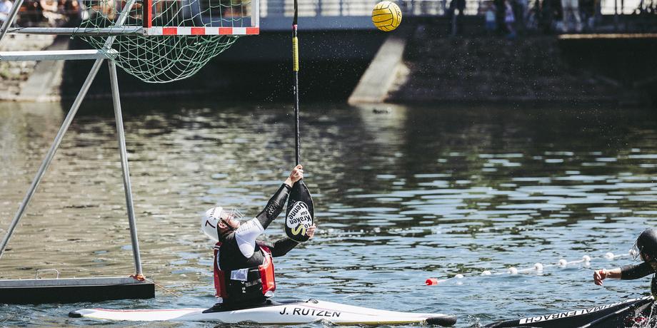 Kajakspieler in Aktion, hebt den Schläger, um einen Wasserball ins Netz zu werfen, während er auf einem Gewässer paddelt.