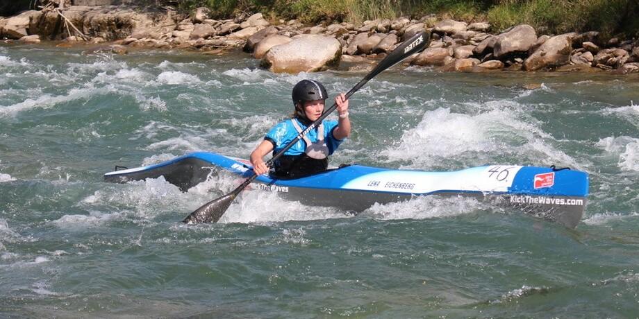 Paddler in einem blauen Kajak manövriert durch rauschendes Wasser, umgeben von Felsen und grüner Vegetation.