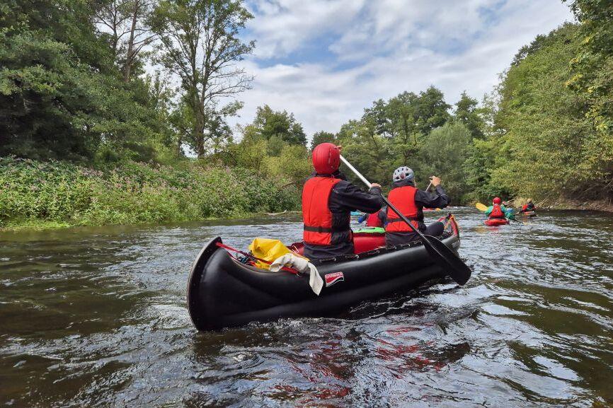 Gruppenausflug mit Kanus auf einem ruhigen Fluss, umgeben von Bäumen und grüner Vegetation unter bewölktem Himmel.
