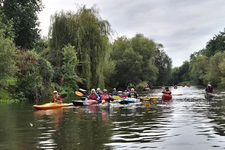 Gruppe von Kajakfahrern auf einem Fluss, umgeben von üppigem Grün und Bäumen unter einem grauen Himmel.