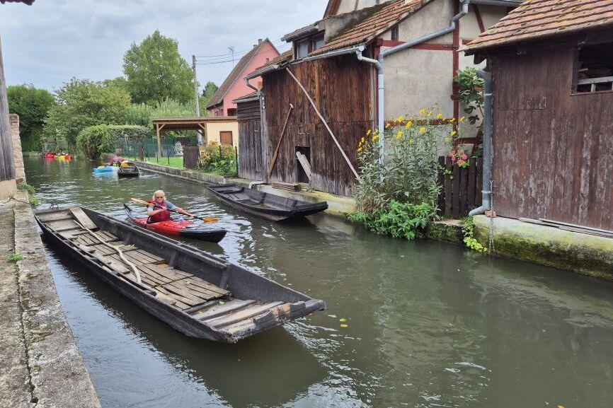 Ruderboot und Kajaks auf einem schmalen Wasserkanal, umgeben von alten Holzgebäuden und Pflanzen.