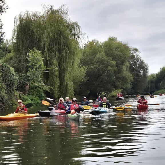 Gruppe von Kajakfahrern auf einem Fluss, umgeben von üppigem Grün und Bäumen unter einem grauen Himmel.
