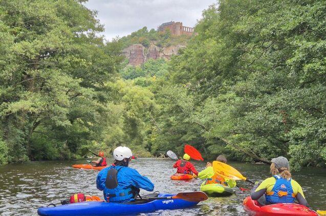 Gruppe von Kajakfahrern auf einem ruhigen Fluss mit üppiger Vegetation und einer Burgruine im Hintergrund.
