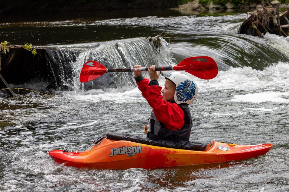 Junge im roten Kajak paddelt mit zwei Paddeln in einem fließenden Gewässer neben einem kleinen Wasserfall.