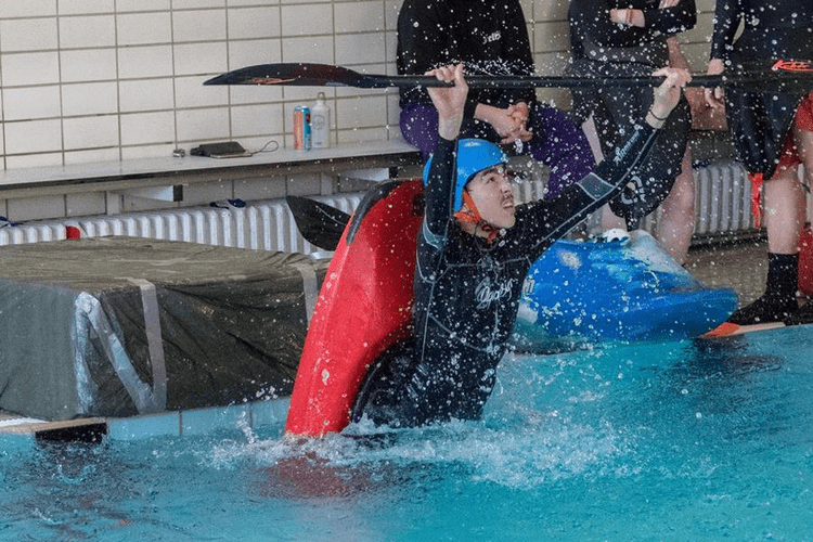 Paddler im Kanu befreit sich beim Training aus einer Kenterübung in einem Hallenbad, umgeben von Zuschauern.