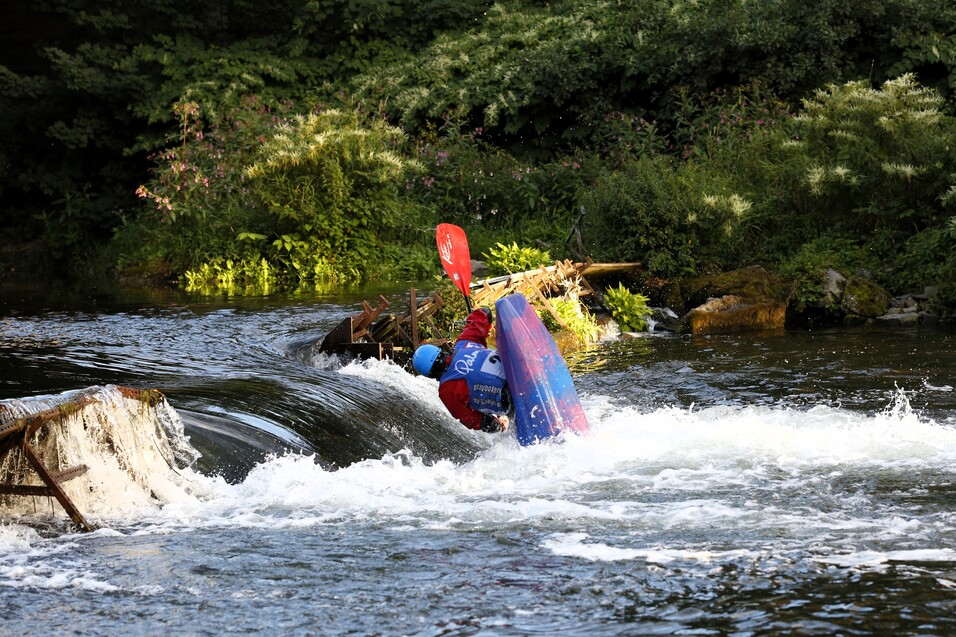 Kajakfahrer in einem türkisfarbenen Kajak, der über eine kleine Wasser oberfläche fährt und spritzende Wellen erzeugt.