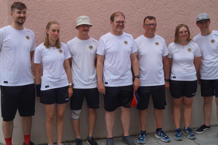 Gruppe von sieben Personen in weißen T-Shirts und sportlicher Kleidung posiert vor einer rosafarbenen Wand.