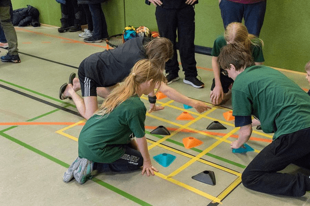 Kinder in grünen T-Shirts spielen ein strategisches Spiel mit bunten Pyramiden auf einem Sportboden.