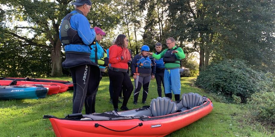 Gruppenschulung zum Kajakfahren: Fünf Personen in Schwimmwesten überprüfen gemeinsam Ausrüstung neben einem roten Kajak.