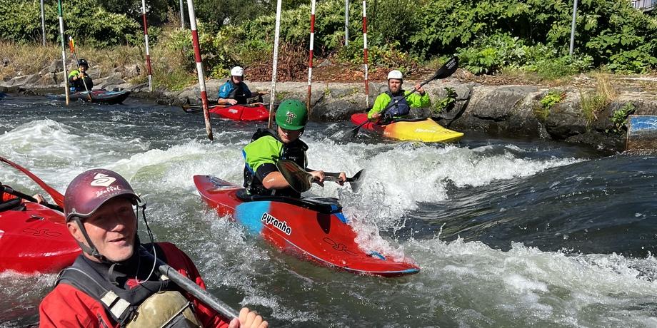 Gruppe von Kajakfahrern auf einem Wildwasser-Kurs, umgeben von Fluss und grüner Vegetation unter blauem Himmel.