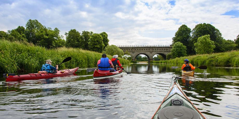 Vier Kajakfahrer paddeln auf einem ruhigen Wasserweg unterhalb einer steinernen Brücke, umgeben von grüner Landschaft.