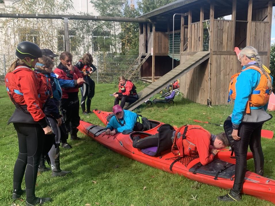 Gruppe von Personen in Schwimmwesten mit Kajak auf der Wiese, im Hintergrund ein Spielplatz.