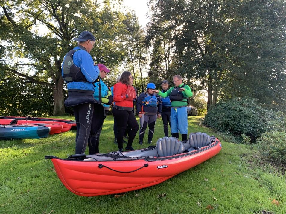 Gruppenschulung zum Kajakfahren: Fünf Personen in Schwimmwesten überprüfen gemeinsam Ausrüstung neben einem roten Kajak.