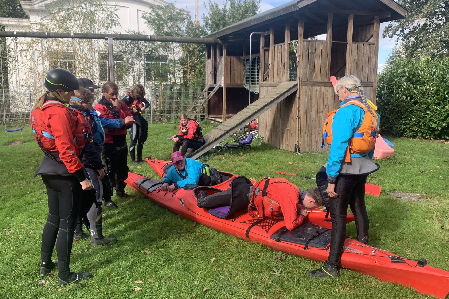 Gruppe von Personen in Schwimmwesten mit Kajak auf der Wiese, im Hintergrund ein Spielplatz.
