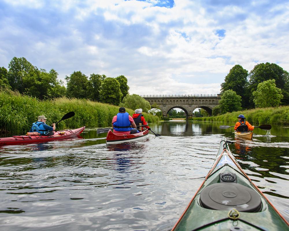 Vier Kajakfahrer paddeln auf einem ruhigen Wasserweg unterhalb einer steinernen Brücke, umgeben von grüner Landschaft.