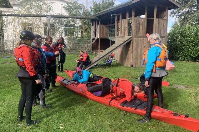 Gruppe von Personen in Schwimmwesten mit Kajak auf der Wiese, im Hintergrund ein Spielplatz.