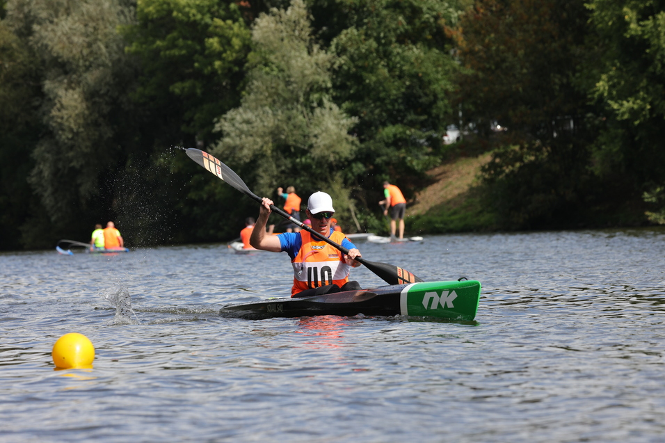 Kanuträger paddelt auf einem Gewässer, während andere Sportler im Hintergrund aktiv sind. Ein gelber Boje schwimmt nahebei.