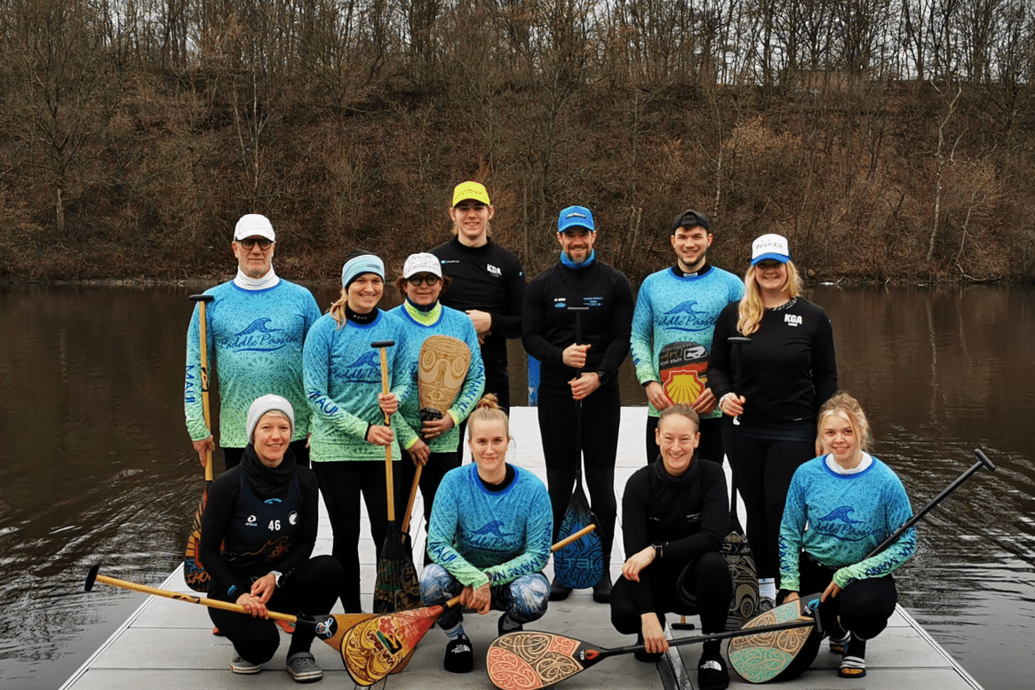 Das Bild zeigt Sportler*innen. Teilweise halten sie Paddel in der Hand. Sie stehen und hocken auf einem Steg. Im Hintergrund sind Wasser und B&auml;ume zu sehen.