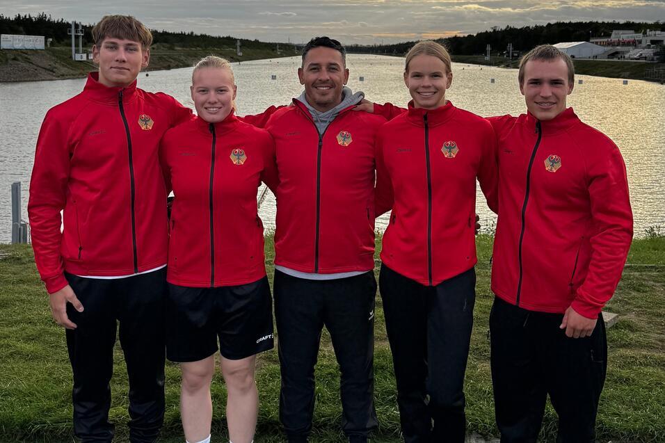 Fünf Personen in roten Jacken stehen vor einem Gewässer unter bewölktem Himmel, mit Bäumen und Gebäuden im Hintergrund.