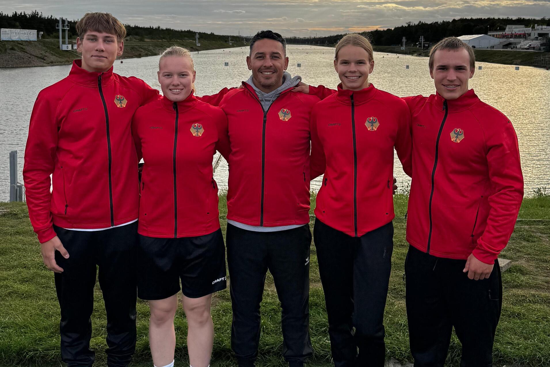 Fünf Personen in roten Jacken stehen vor einem Gewässer unter bewölktem Himmel, mit Bäumen und Gebäuden im Hintergrund.