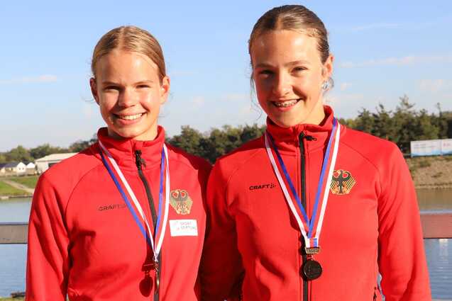 Zwei Sportlerinnen in roten Jacken mit Medaillen lächeln vor einem Gewässer in der Sonne.