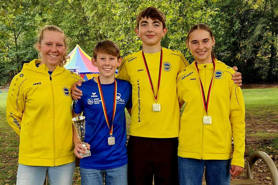Vier junge Sportler in gelben Trainingsjacken stehen lächelnd auf einer Bank im Freien, halten Medaillen und einen Pokal.