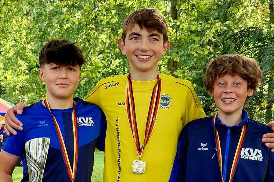 Drei Jugendliche mit Medaillen und Pokal stehen lächelnd in einem Park unter Bäumen.