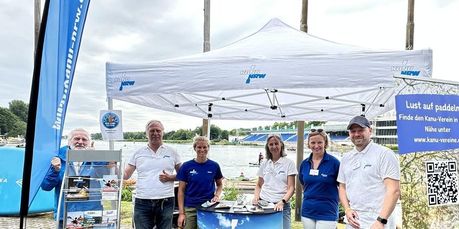 Sechs Personen stehen vor einem Informationsstand zum Thema Kanu in NRW, umgeben von Wasser und Pflanzen.