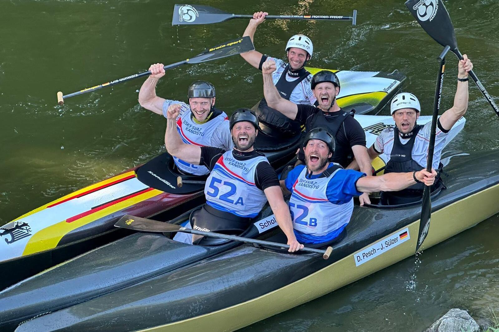 Sechs männliche Kanuten feiern in Booten auf einem Fluss, paddeln und heben die Arme in der Luft.