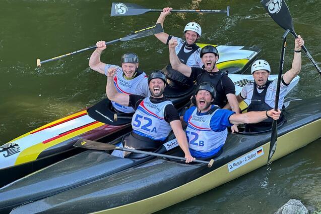 Sechs männliche Kanuten feiern in Booten auf einem Fluss, paddeln und heben die Arme in der Luft.