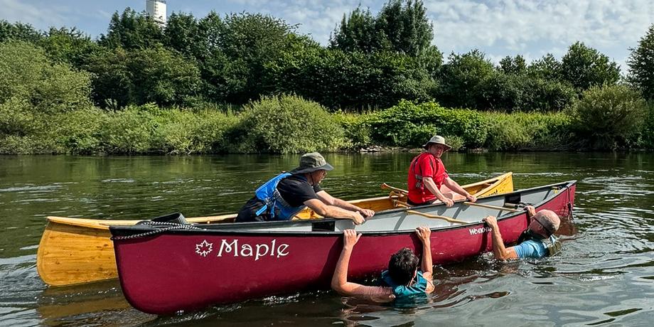 Personen in Kanus auf einem Gewässer, einer fällt ins Wasser, umgeben von grüner Vegetation und bewölktem Himmel.