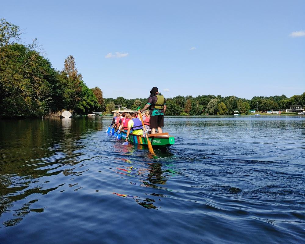 Personen in einem Boot paddeln auf einem ruhigen Gewässer unter blauem Himmel mit Bäumen im Hintergrund.