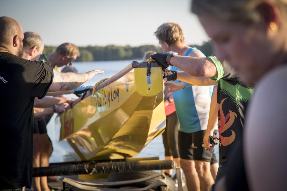 Mehrere Personen heben gemeinsam ein gelbes Drachenboot an einem Gewässer in einer sonnigen Umgebung.