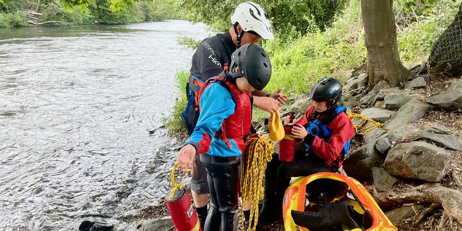 Drei Personen in Neoprenanzügen bereiten sich am Ufer eines Flusses mit einem Kanu und Rettungsgeräten vor.