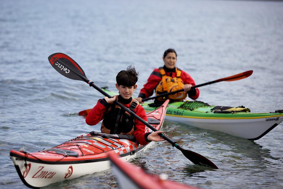 Zwei Personen in Kajaks paddeln auf ruhig fließendem Wasser, eine in rotem und die andere in grünem Kajak.