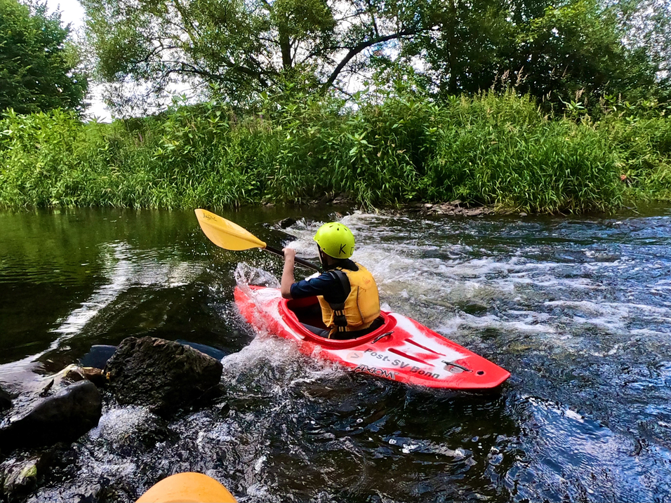Junge im roten Kajak paddelt auf einem ruhigen Gewässer, umgeben von grünen Pflanzen und Bäumen.