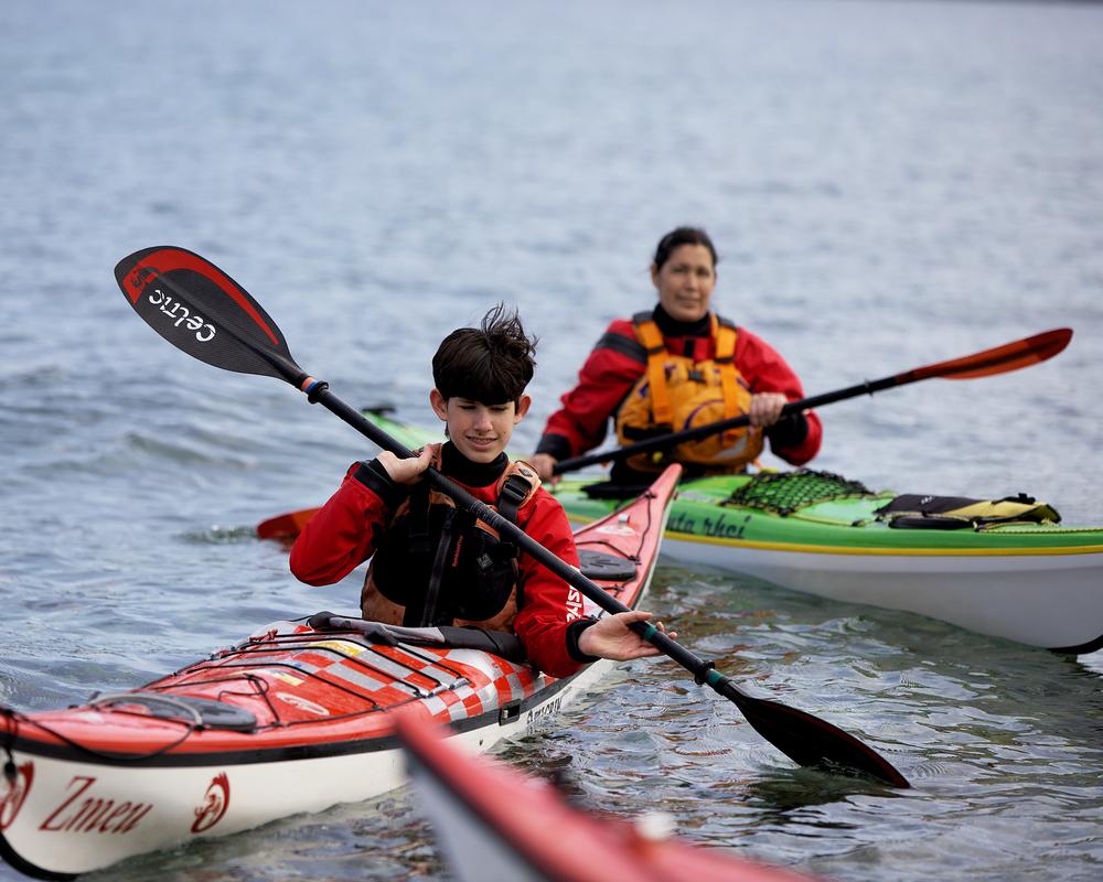 Zwei Personen in Kajaks paddeln auf ruhig fließendem Wasser, eine in rotem und die andere in grünem Kajak.