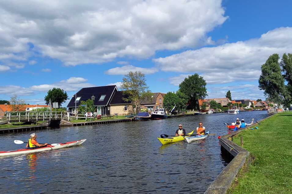 Kanufahrer paddeln auf einem Kanal, umgeben von grüner Wiese und Häusern unter einem blauen Himmel mit Wolken.