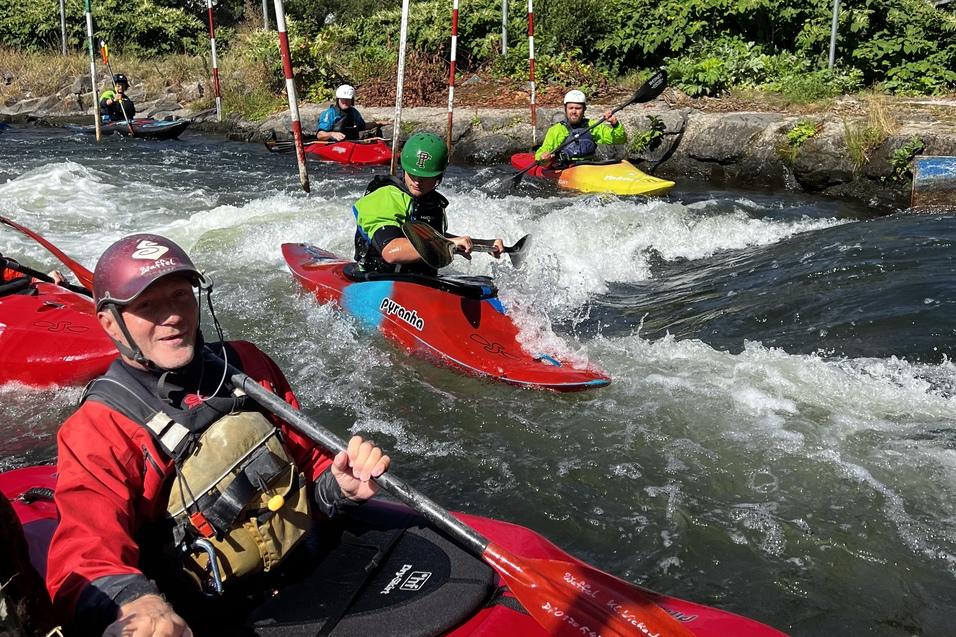 Kaiakfahrer paddeln in rotem Kajak durch eine Welle an einer Wildwasserstrecke mit grüner Umgebung.