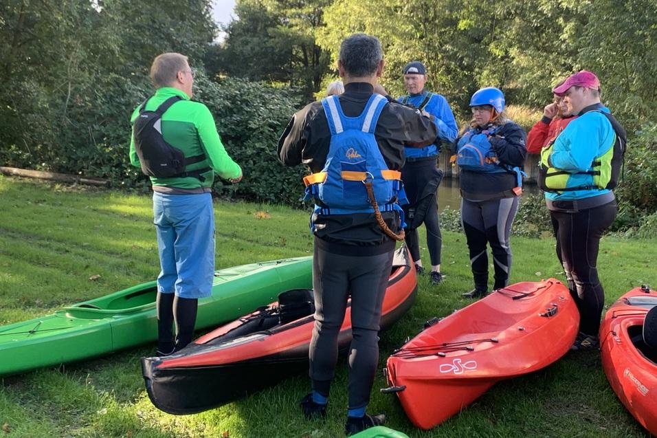 Gruppe von Kanufahrern in Neoprenanzügen und Schwimmwesten bespricht paddeln vor roten und grünen Kajaks auf dem Rasen.