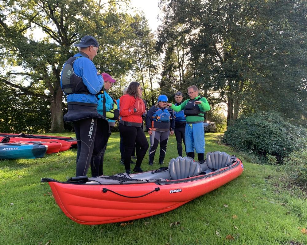 Sechs Personen in Paddeljacken und Schwimmwesten besprechen gemeinsam eine rote Kajak/kanu auf einer Wiese.