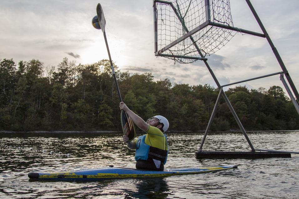 Mann in einem Kajak hebt den Paddel, um einen Ball in ein hohes Netz über Wasser zu werfen. Hintergrund mit Bäumen.