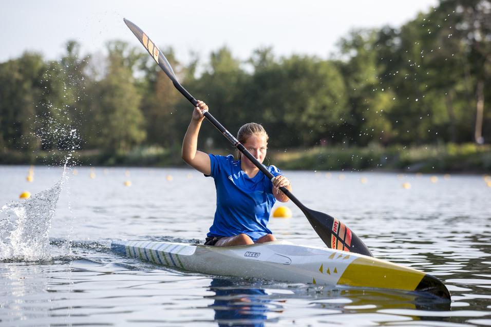 Sportlerin paddelt in einem Kanu auf einem See, mit Spritzern im Wasser und grüner Ufervegetation im Hintergrund.