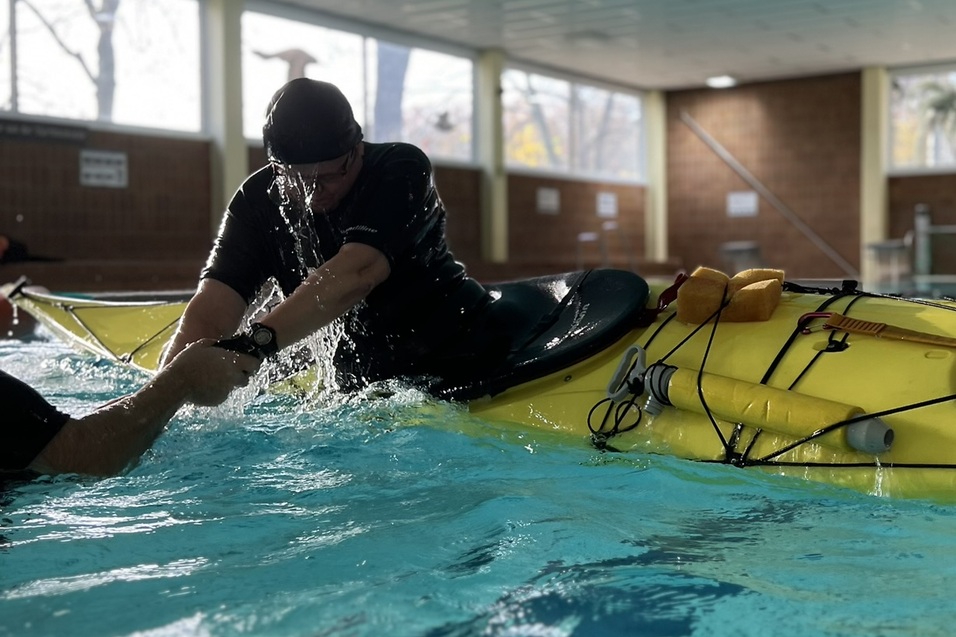 Person in einem Kanu im Wasser greift nach einer anderen Person, während Wasser spritzt. Hallenbad im Hintergrund.