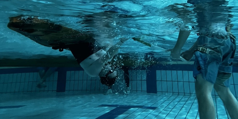 Person unter Wasser mit Schwimmbrille, während eine andere Person am Schwimmbeckenrand einen Schwimmring hält.