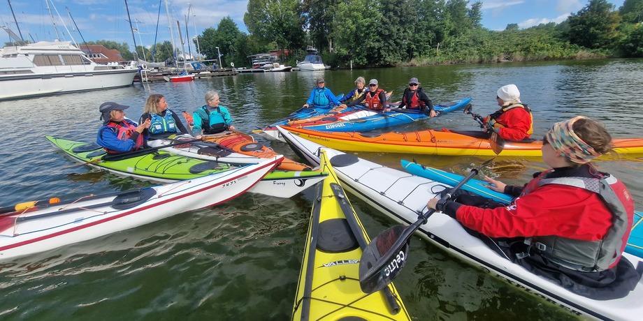 Gruppe von Kajakfahrern in bunten Kajaks versammelt sich auf Gewässer nahe einem Yachthafen unter blauem Himmel.