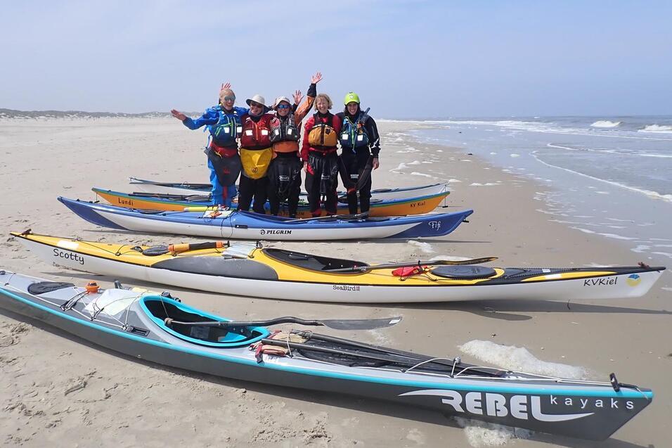 Gruppe von Kajakfahrern steht auf Kajaks am Strand; Wellen im Hintergrund, bewölkter Himmel.